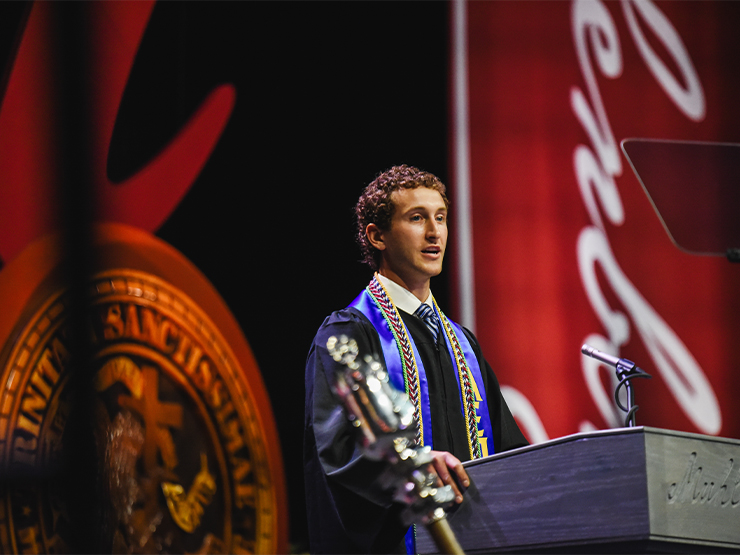 A student wearing graduation regalia speaks at a podium. Muhlenberg banners and crest take up the stage behind him.