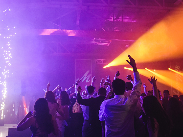 A room full of dressed up young adults raise their hands and dance in a room filled with dramatic purple and gold concert lighting.