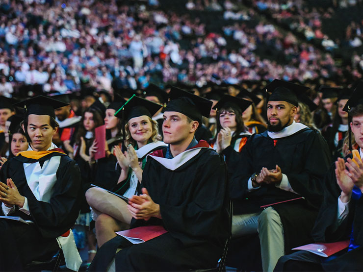 Applauding young adults dressed in graduation caps and gowns fill the floor of an arena.