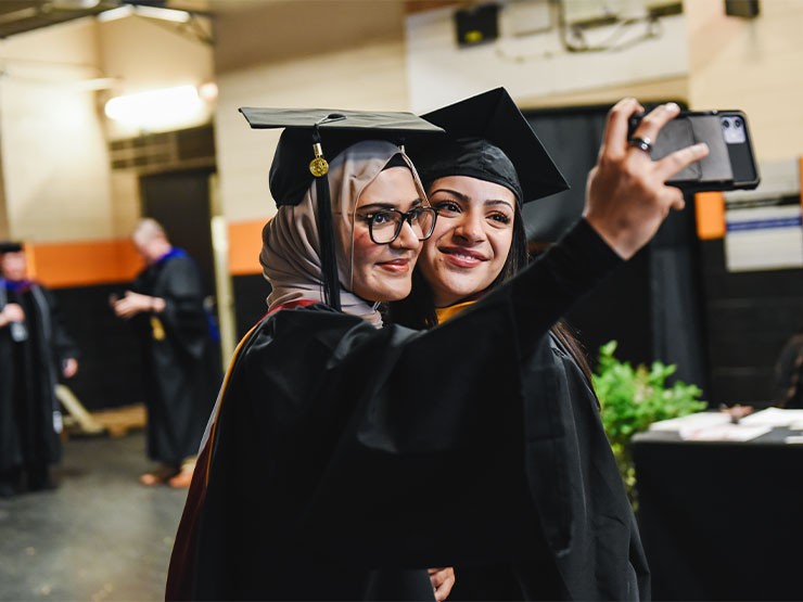 A pair of young adults dressed in commencement regalia stand close to one another and pose for a cell phone selfie.