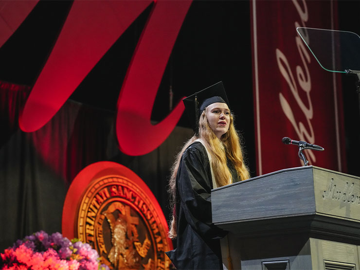 A college student with long blonde hair and dressed in graduation regalia speaks at a podium engraved with 