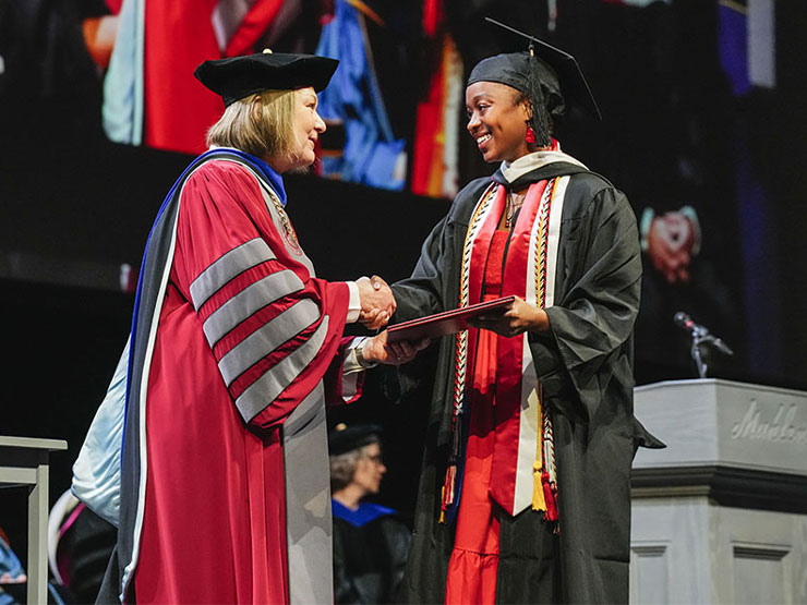 A woman dressed in college presidential regalia shakes hands and offers a diploma to a smiling graduate.