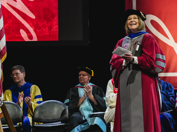 A woman in crimson college president regalia smiles and claps while standing on stage and looking out over an audience.