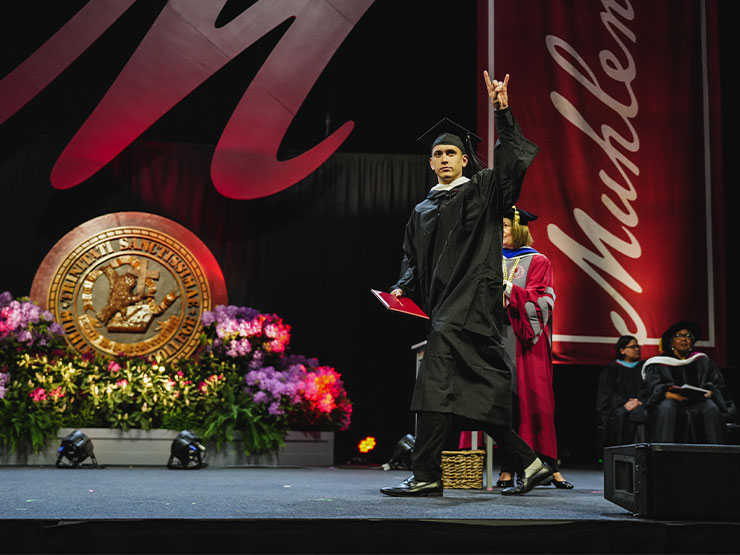 A college student in graduation regalia walks across a stage, one hand holding a diploma and the other aloft in a 