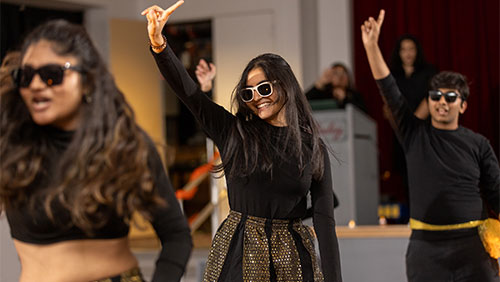 Students in black and gold dress dance while wearing sunglasses at a Diwali event.