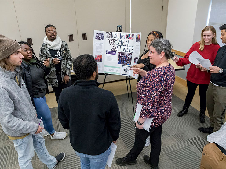 An adult speaks to a group of young adults over a poster titled 