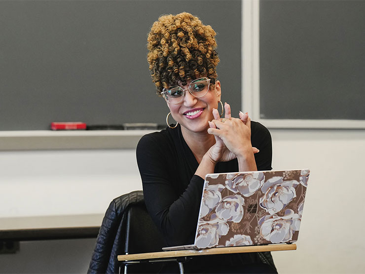 A college professor sits in front of her class with a laptop open on the desk in front of her