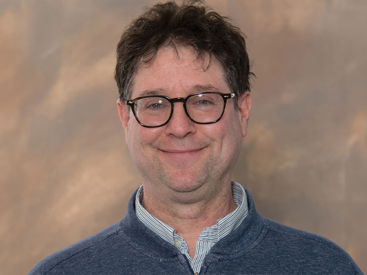 A college professor with glasses smiles at the camera against a beige background.