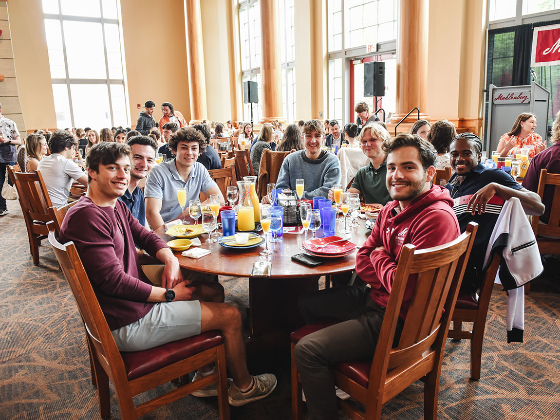 A group of smiling college students gather around a table in a dining hall