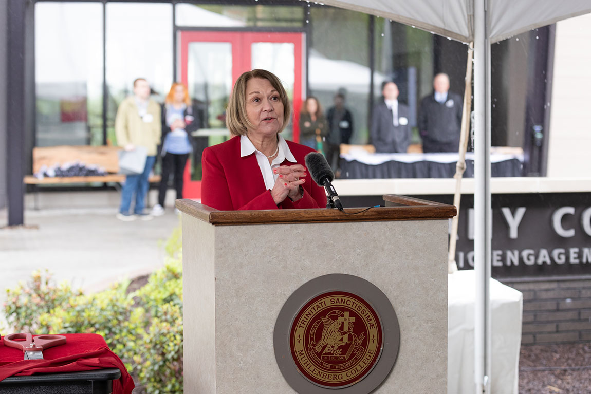 A woman in a red blazer and white shirt speaks at a podium outside in front of a new building with red doors