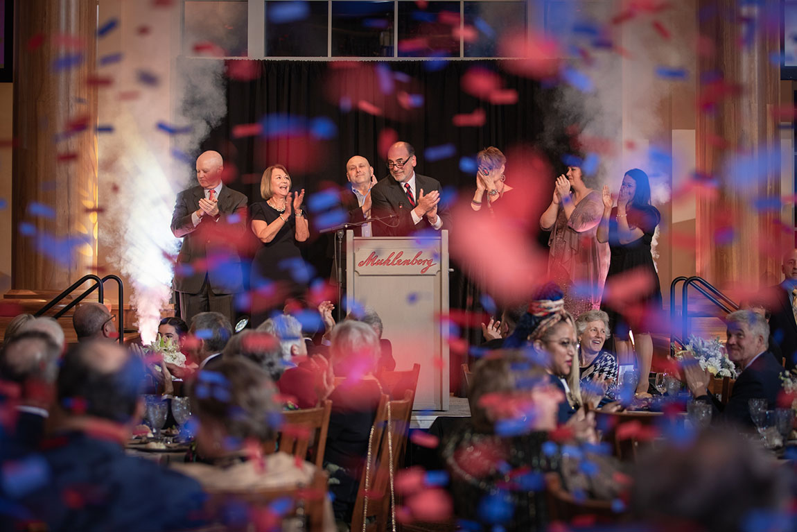 A group of people applaud on a stage in front of a crowd as blue and red confetti rains down