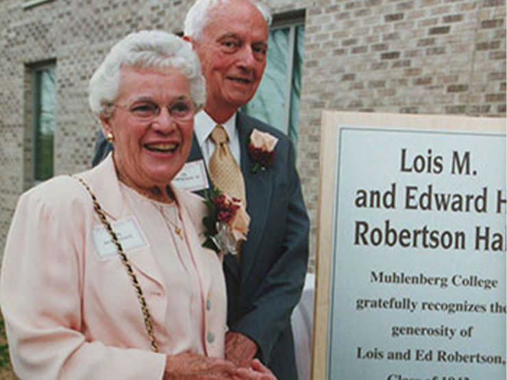 Two older adults smile at the camera by a plaque commemorating a donation.