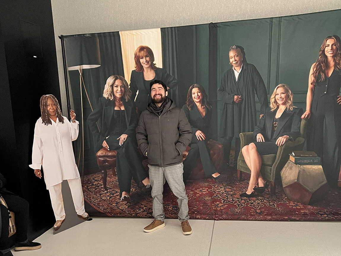 A college student poses in front of a backdrop with several women on it, the cohosts of The View