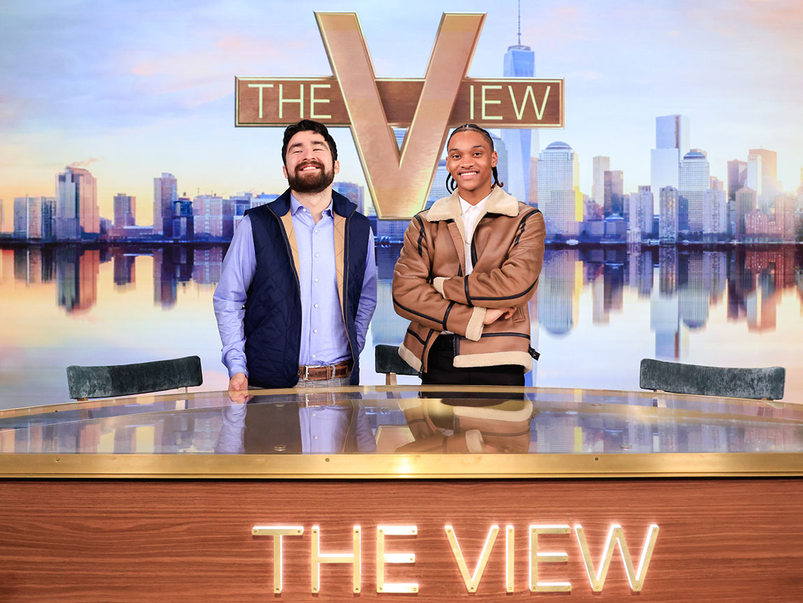 Two college students pose behind a desk that says THE VIEW