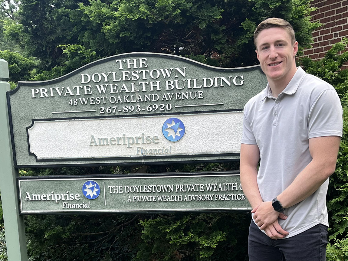 A young person in a gray shirt stands next to a sign that says THE DOYLESTOWN PRIVATE WEALTH BUILDING - Ameriprise Financial