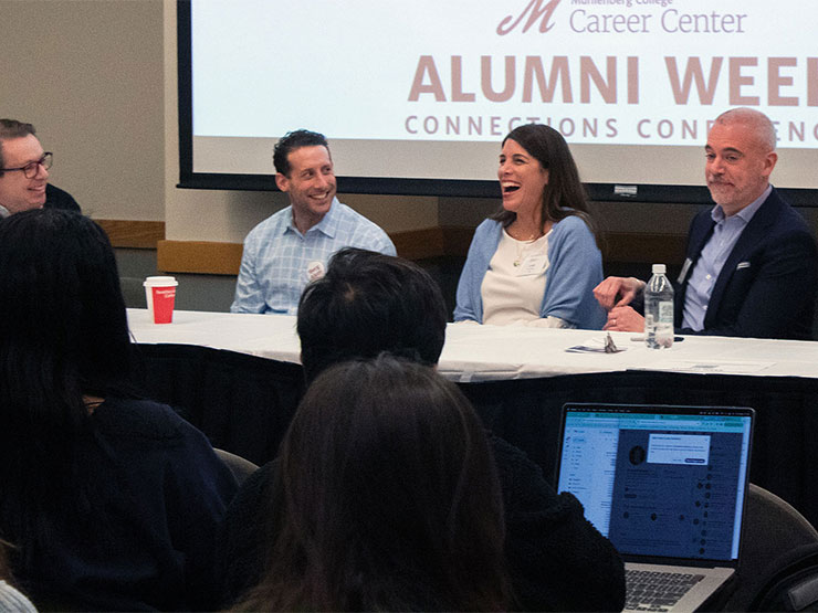 Four people on a panel sit at a table in front of a group of students.