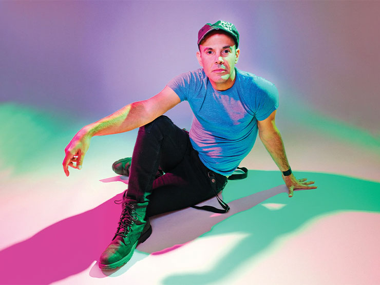 A  man poses on the ground, hand over a bent knee, in a room with colorful lighting.
