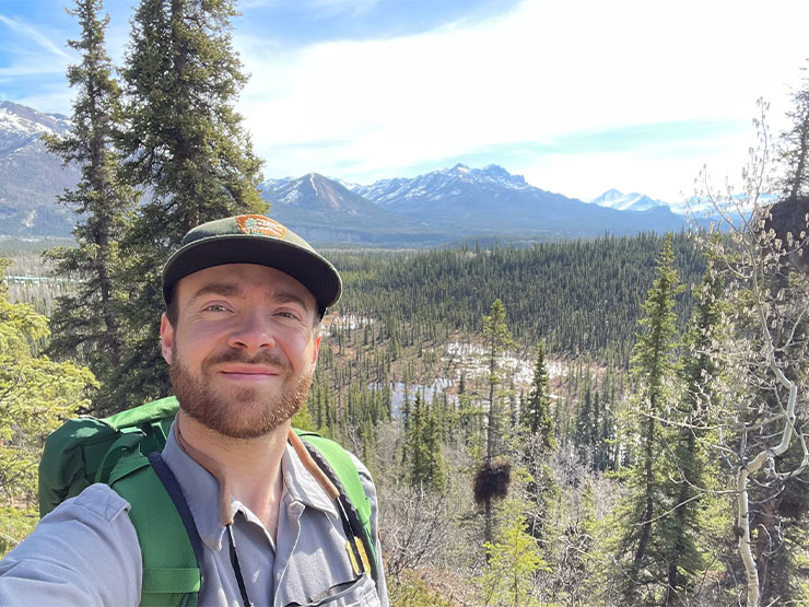 A young adult in a hat with a beard takes a selfie in the forest with snow-capped mountains in the background