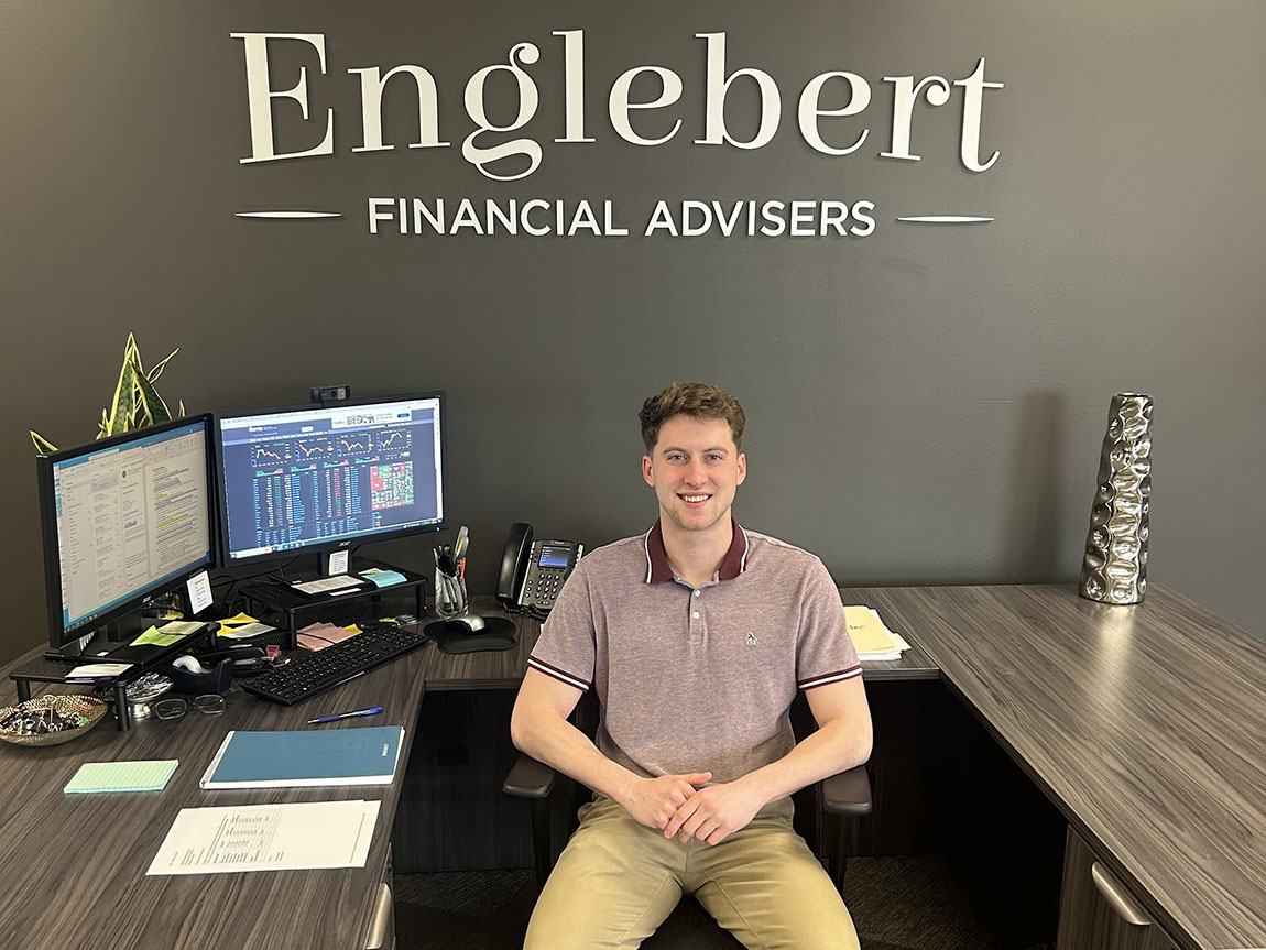 A college student in business casual clothes sits at a computer and smiles at the camera. The wall behind him reads Englebert Financial Advisers