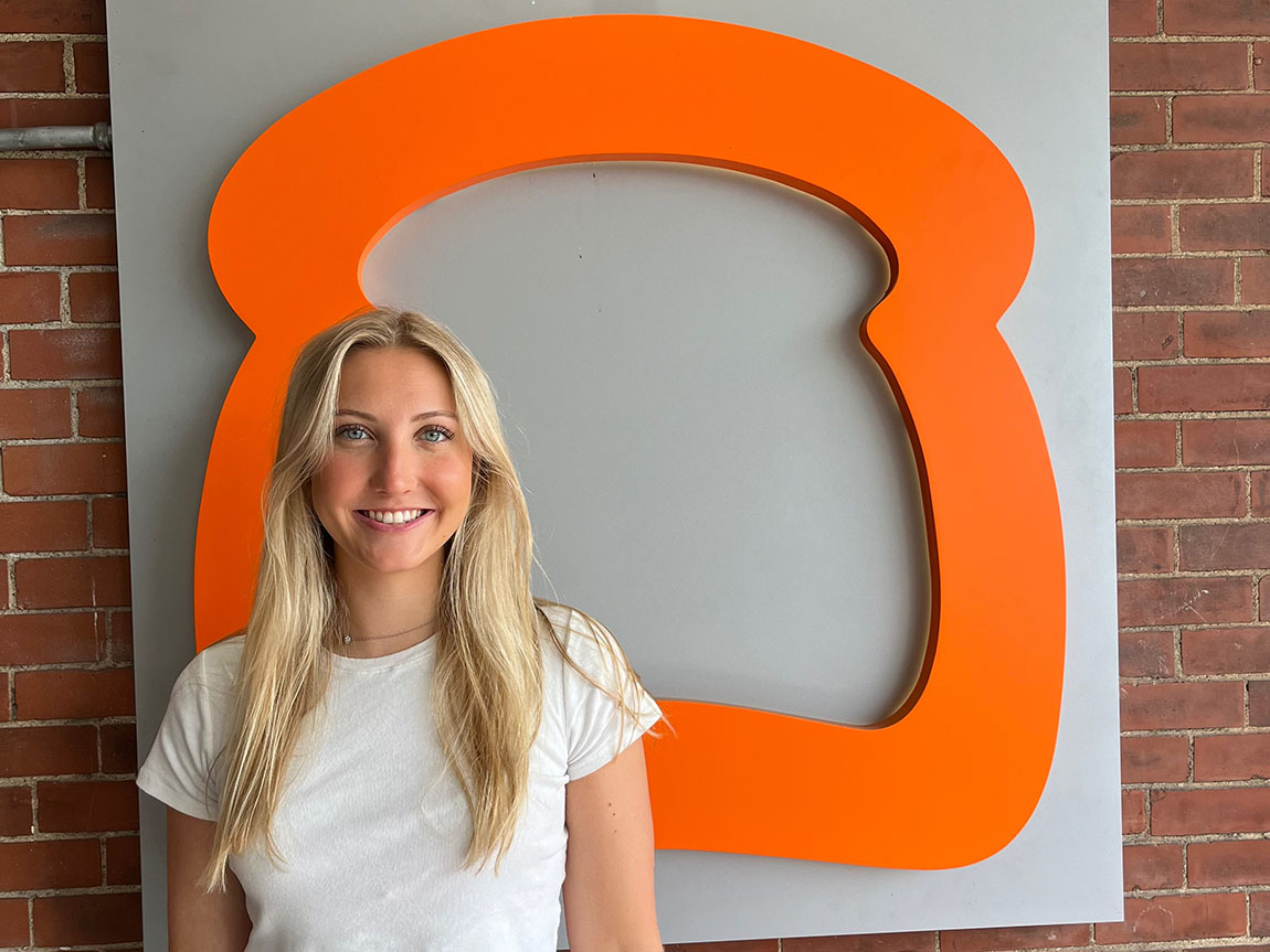 A young woman smiles for a photo in front of an orange outline that looks like the shape of a piece of toast