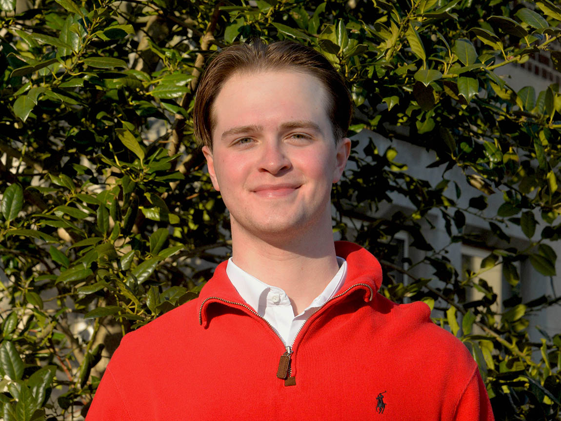 A college student in a red sweater stands in front of a wall covered in green leaves