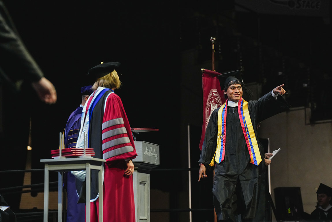 A college graduate points at the crowd while walking across the stage to receive a diploma