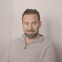 A headshot of a college professor with facial hair wearing a tan shirt