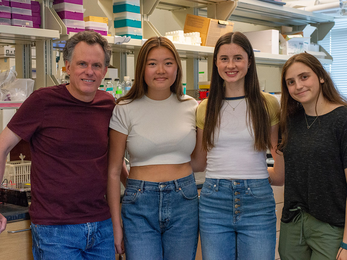 A college professor stands next to three college students in a science lab