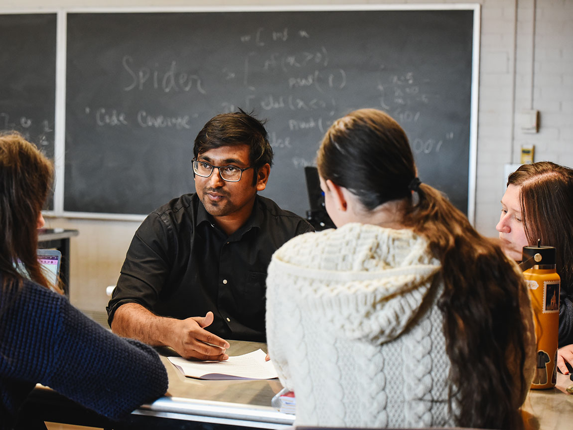 A college professor sits at a table and explains something to students with a chalkboard in the background