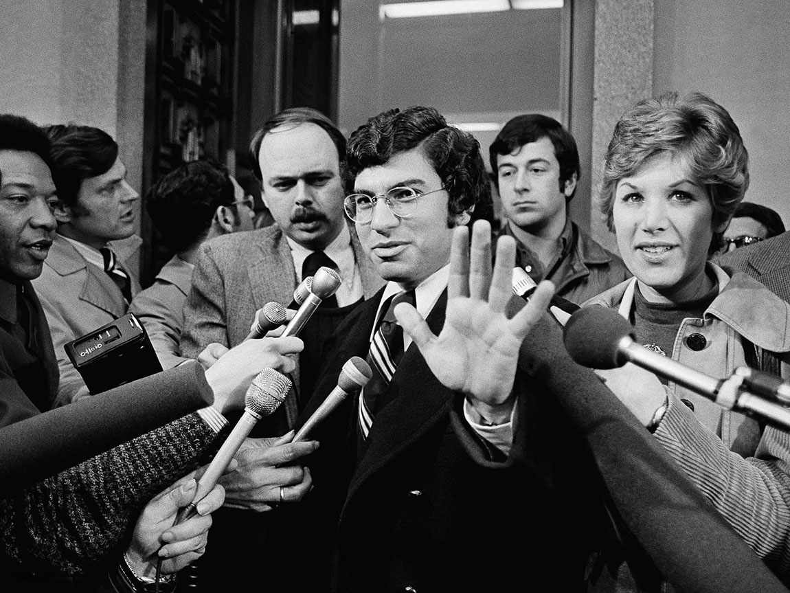 An archival black and white photo of a man in glasses raising a hand toward reporters' microphones during the Watergate scandal