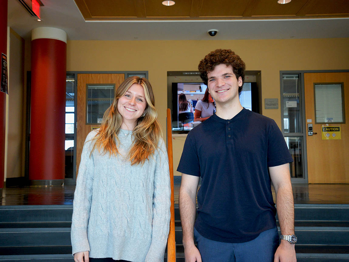 Two smiling college students stand next to each other in the lobby of a science building