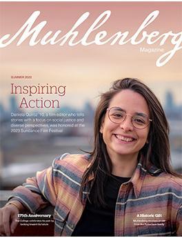 On the cover of a magazine with the cursive script Muhlenberg, an adult with shoulder-length brown hair and glasses smiles at the camera with the city skyline in the distance horizon.