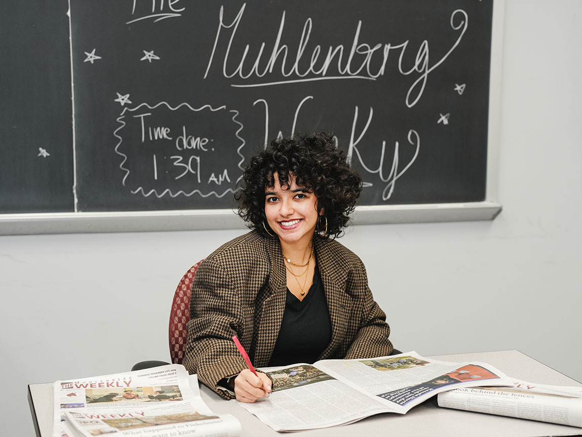 A college student in a blazer sits in front of a chalkboard that says The Muhlenberg Weekly