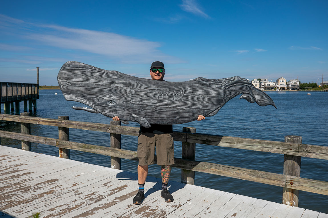A man in a black baseball cap holds a giant wooden carved whale as he stands on a dock with water in the background