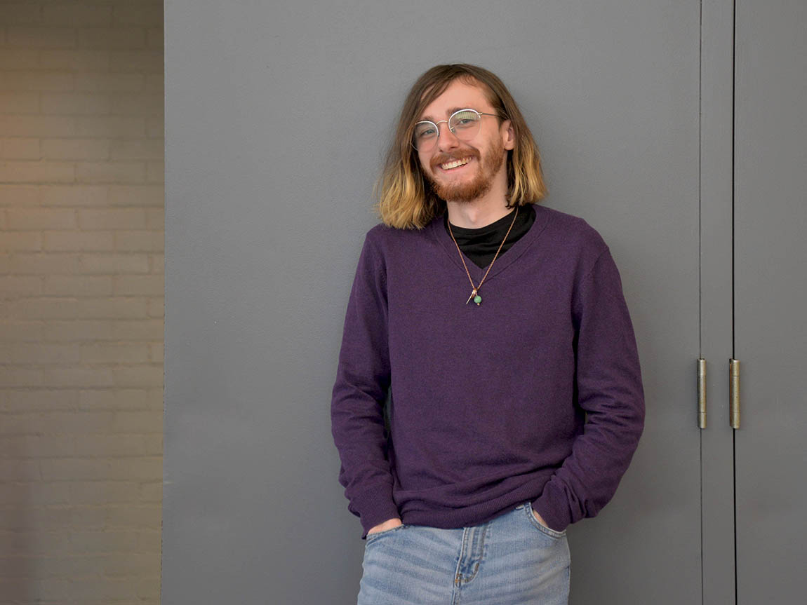 A college student with long hair and glasses wearing a purple sweater poses for a portrait