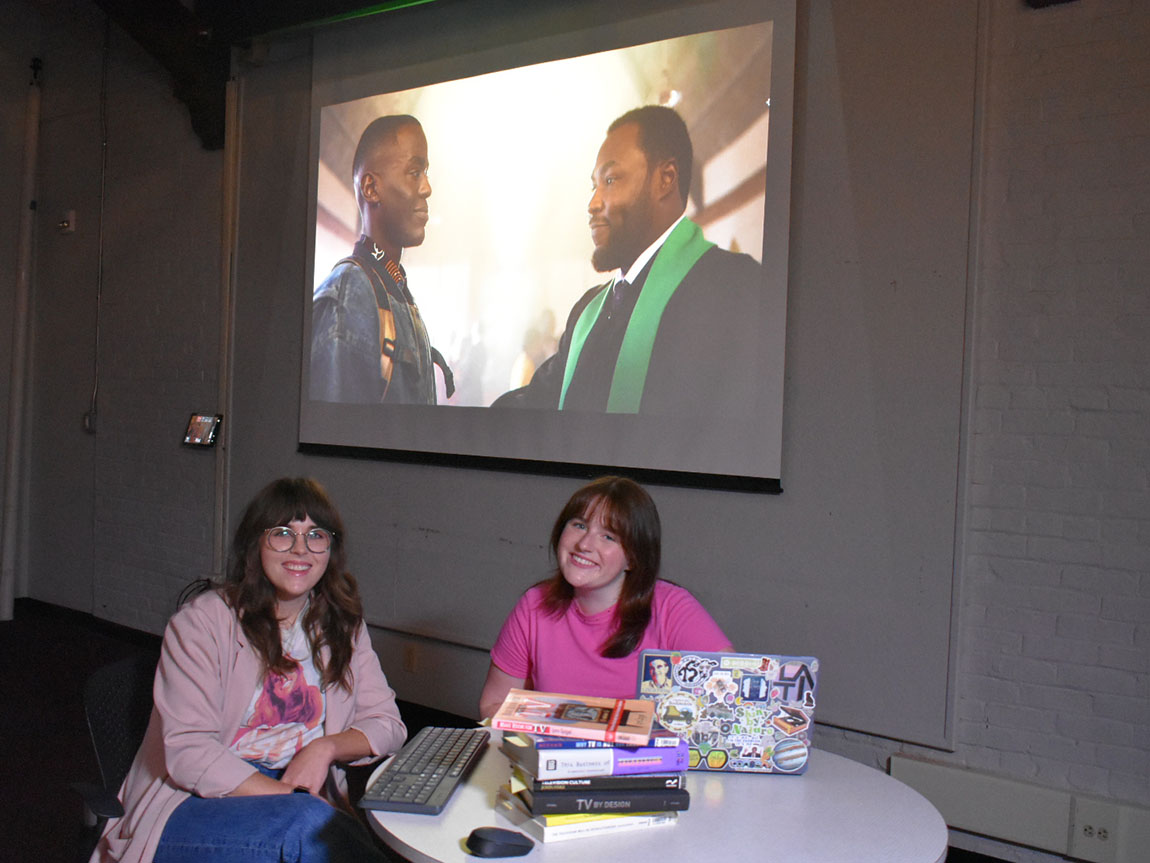 A college professor and college student smile in front of a screen showing a TV show