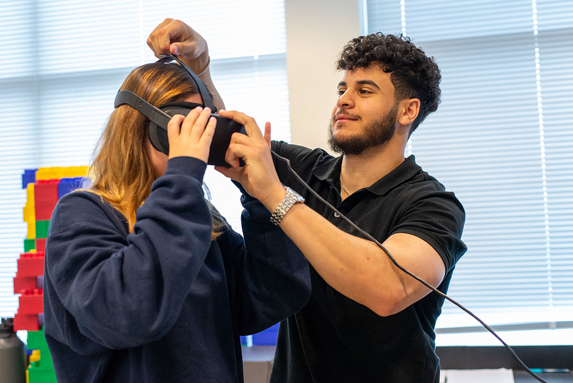 One college student helps another college student put on a VR headset