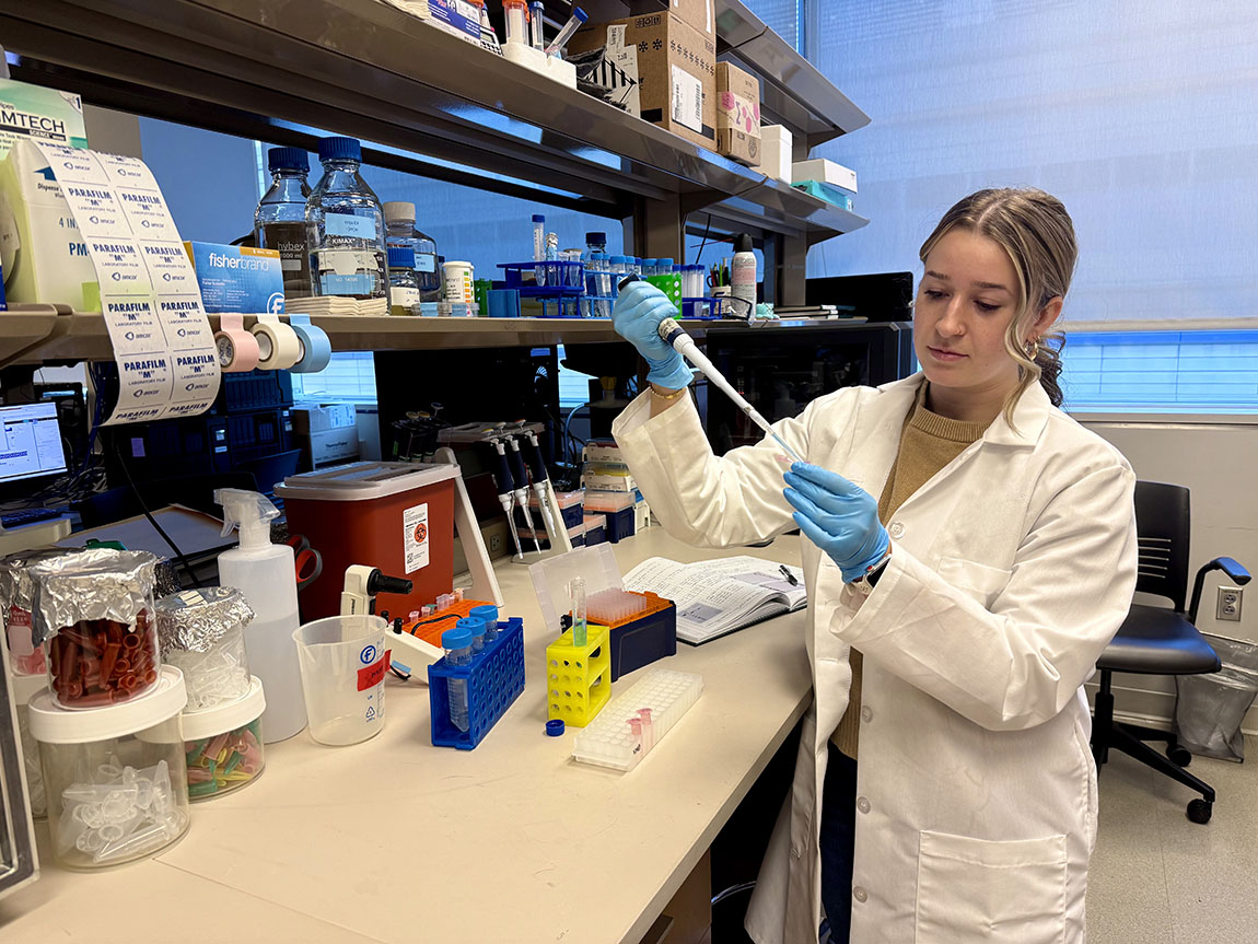 A Ph.D. student in a white lab coat and blue gloves uses a dropper into a test tube