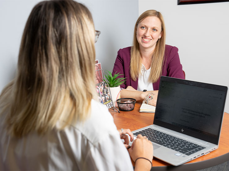 A woman in a maroon blazer sits across a desk with another person with an open laptop in front of them.