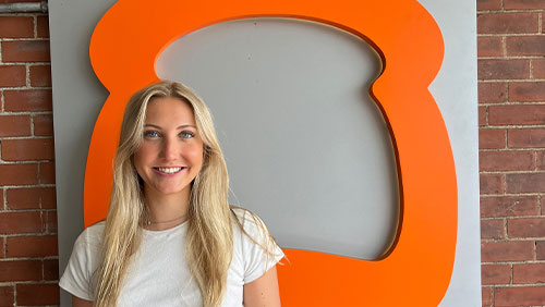 A young adult with long blonde hair smiles while standing next to an orange logo shaped like a piece of toast.