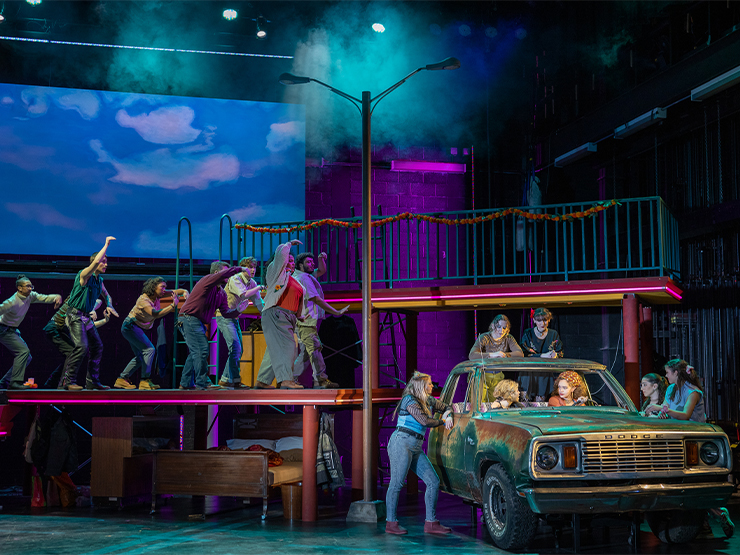 Students in a stage performance gather on a platform and around a rusty car.