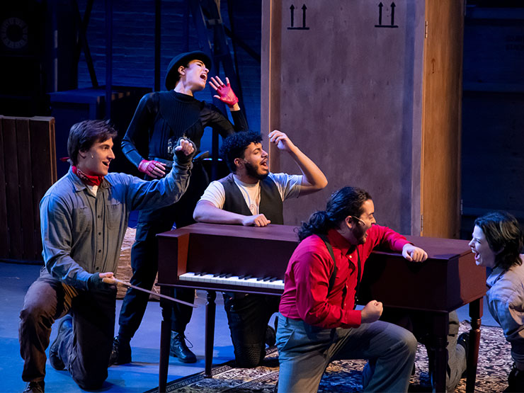 A group of young adults sing around a piano on a stage with sparse scenery.