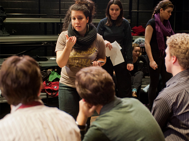 A student, standing, speaks to a group of seated students inside a theatre studio space.