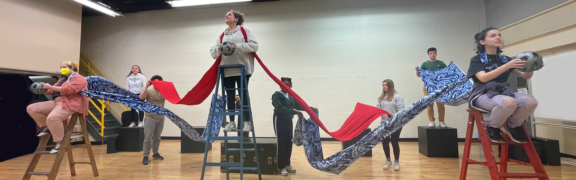 Eight college-age actors work in a rehearsal hall. Several of them are sitting or standing on ladders and wooden blocks, and they are all interconnected by long, flowy pieces of brightly colored fabric, which they are shaking up and down.
