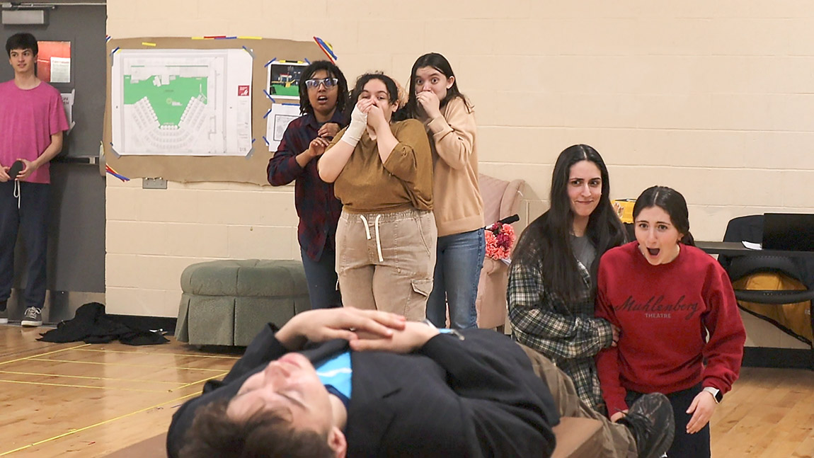 A group of students rehearses a play. In the foreground, an actor lies on his back, hands folded on his chest. Behind him, five other actors express shock.