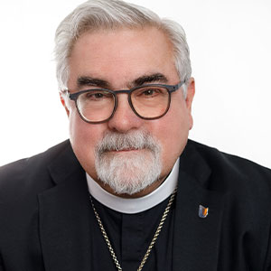 An adult with glasses and white hair wears a black robe with a clerical collar and looks to the camera.